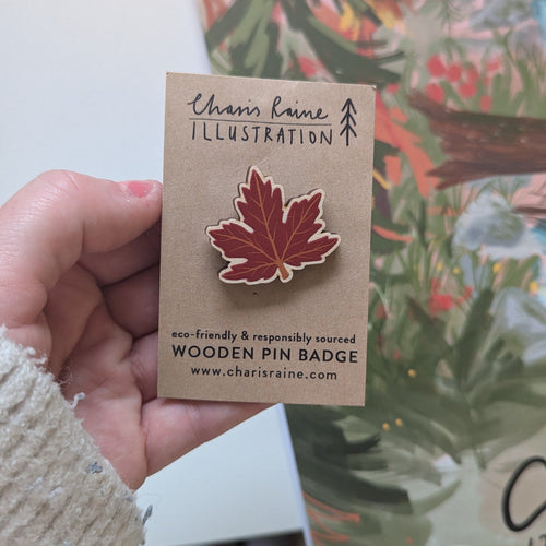 Hand holding a wooden pin badge on a card with 'Charis Raine Illustration' branding.