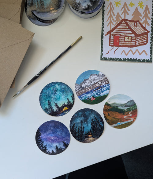 Five round painted artworks on a white surface with a paintbrush and a printed card of a cabin.